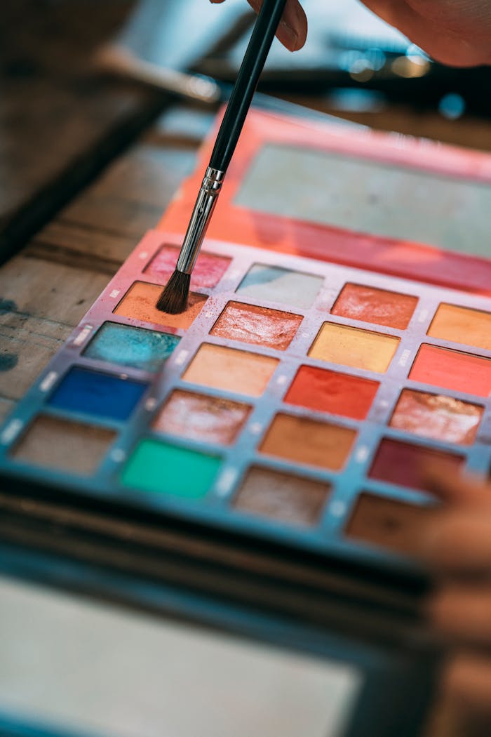 gallery-5 Close-up of a colorful eyeshadow palette with a brush applying makeup, highlighting creativity.